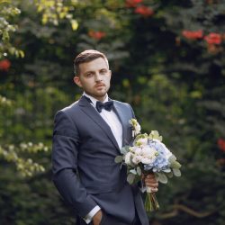 Untitled-8_0001_handsome-stylish-groom-blue-suit-stands-park-near-bushes-with-bouquet